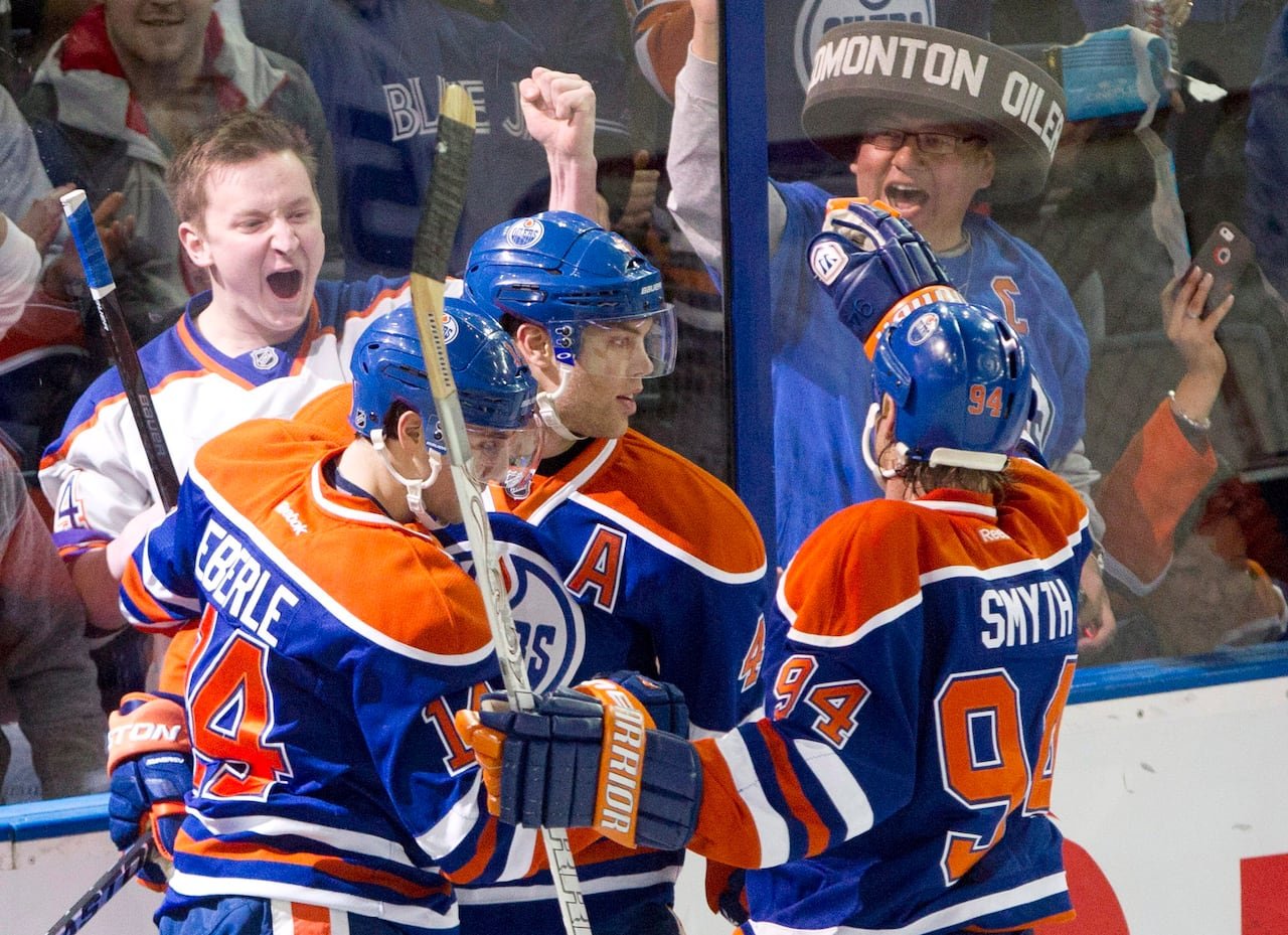 default-359 Three hockey players in orange-and-blue jerseys embrace joyously as fans on the opposing side of the arena glass cheer fervently.
