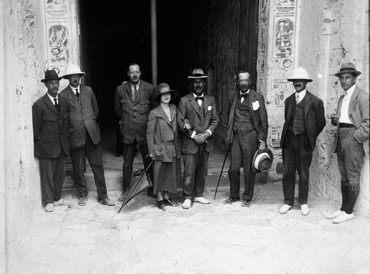 Seven men and one woman stand in front of the entrance of a stone tomb. The photograph is in black and white.